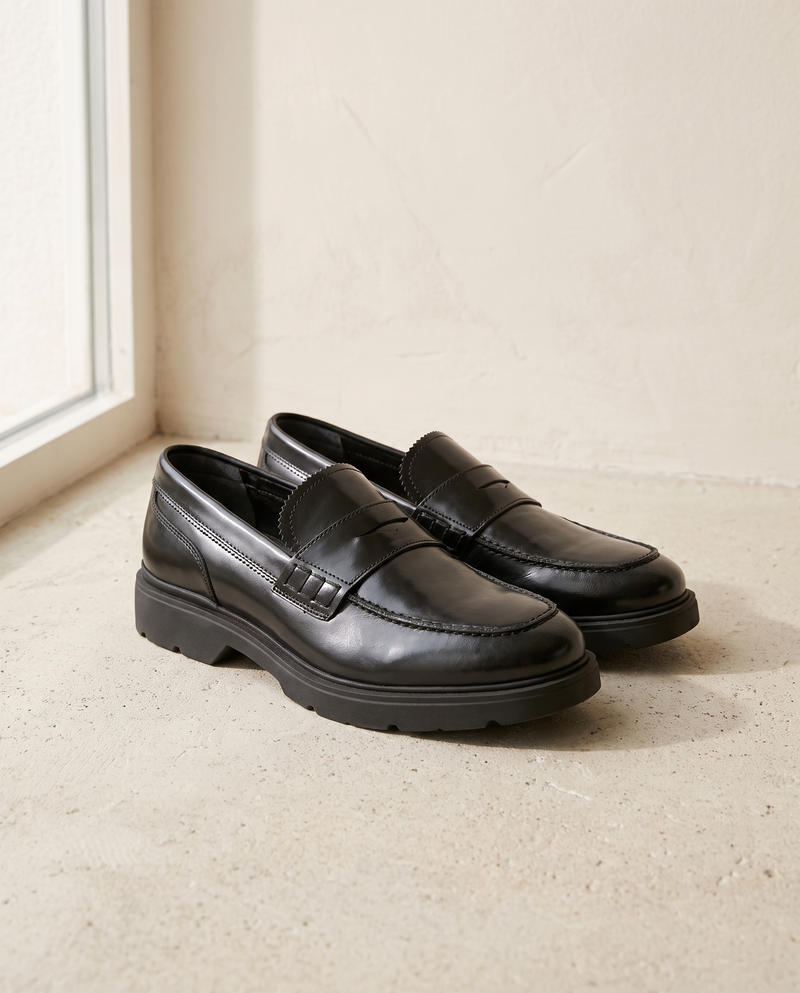 Mocassin ARCHIBALD noir posé sur sol béton clair, mur enduit crème