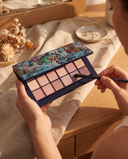 Palette fards à paupières Natural Nude CXL by Christian Lacroix tenue en main avec un pinceau, sur une coiffeuse en bois