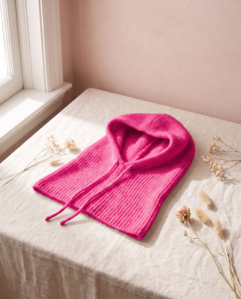 Cagoule maille Primerose fuchsia, posée sur un lin crème, mur rose poudré