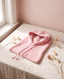 Cagoule maille Primerose rose bonbon, posée sur un lin crème, mur rose poudré