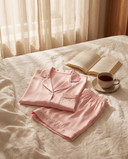 Set de pyjama court en soie Maëlla rose clair plié sur un lit en lin, livre et tasse de thé