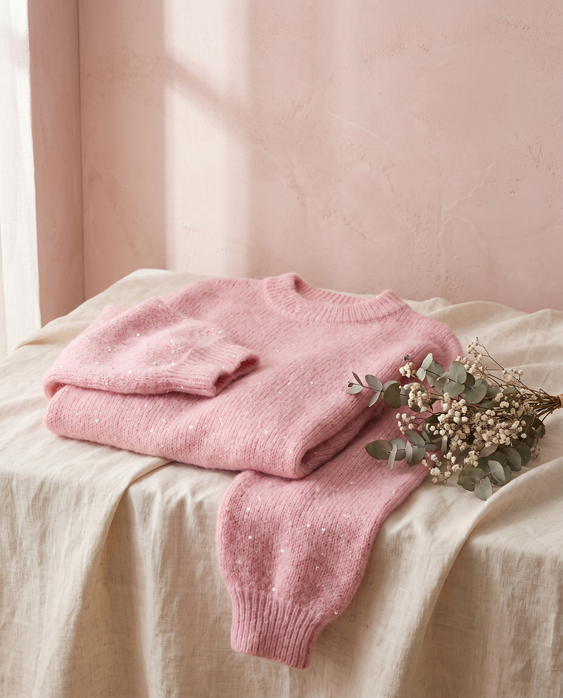 Pull paillettes Solveig rose, posé sur un lin crème, mur rose poudré